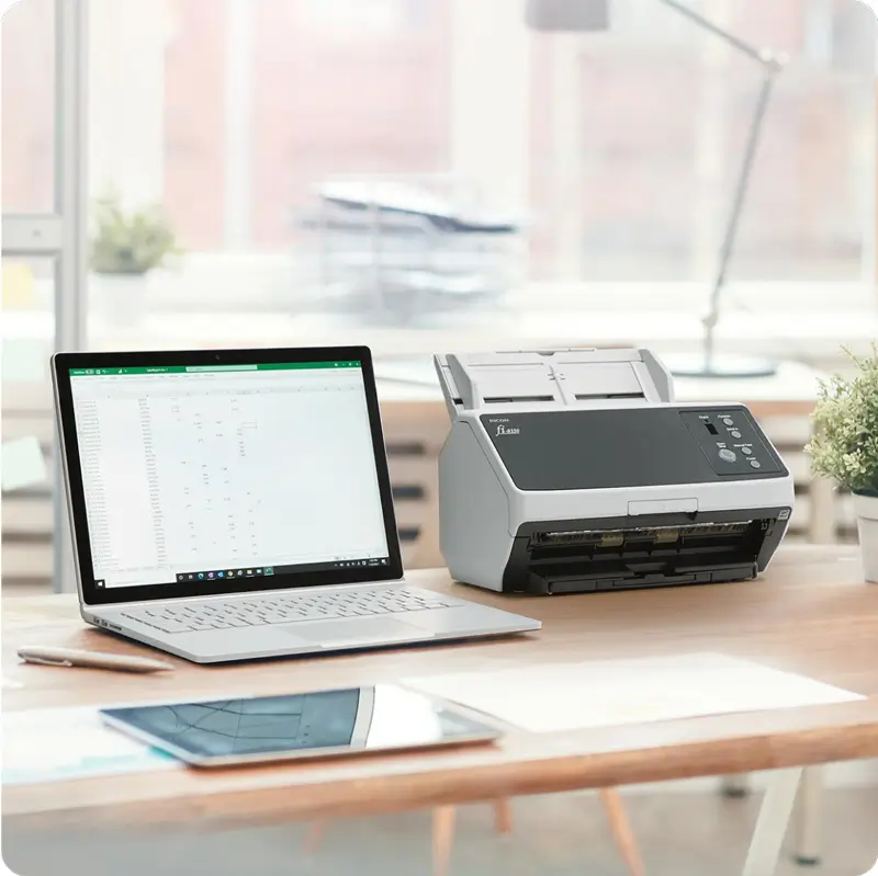 Office setup with a laptop and a high-speed document scanner on a wooden desk, in a bright workspace with a tablet and potted plant nearby.